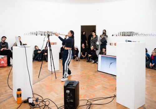 Artist has her mouth to a blowgun and is standing in front of a 3D scanning machine in the middle of an art gallery