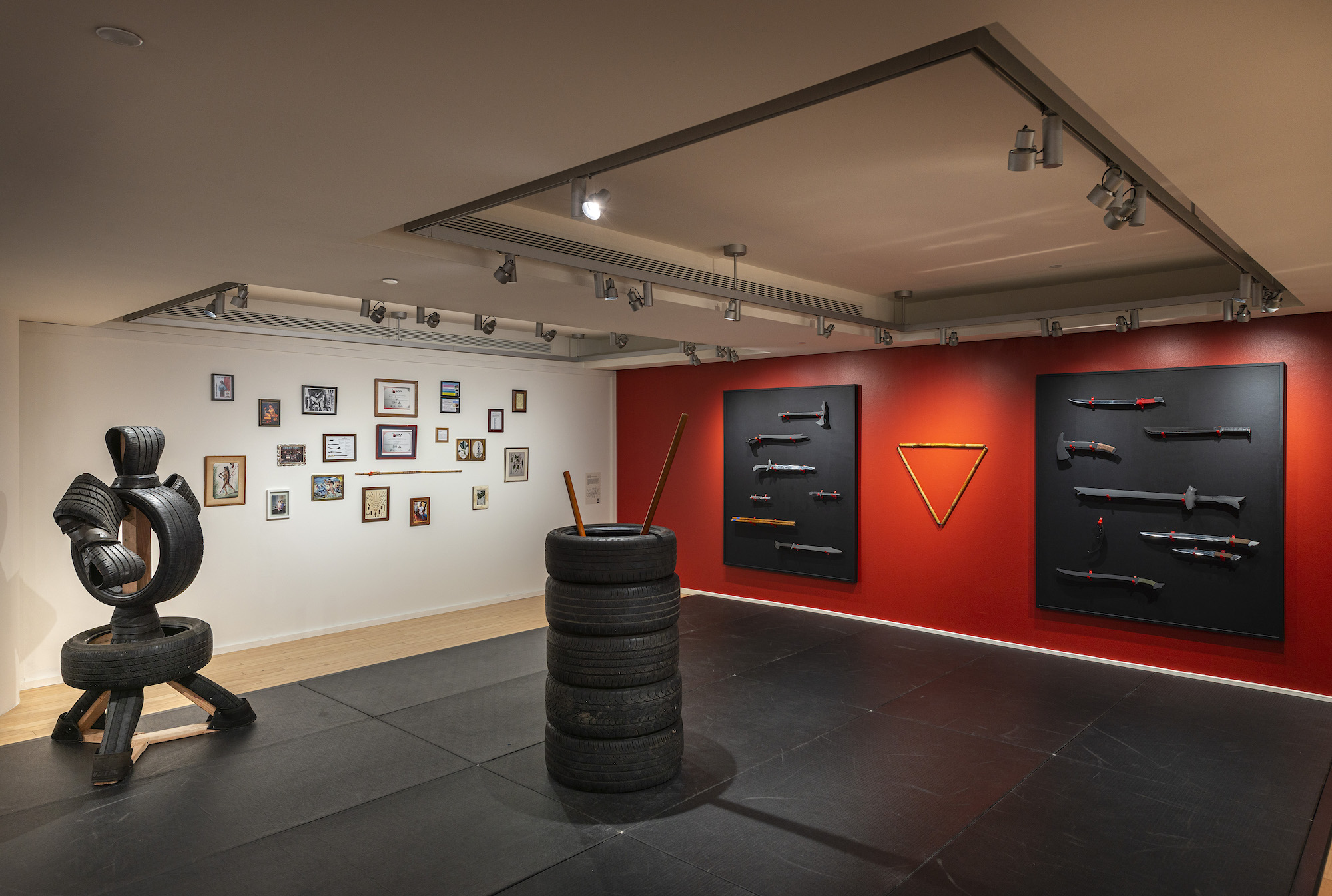 A gallery with a stack of tires in the center, a tire dummy sculpture next to it, as well as training weapons in a red wall and framed images on the adjacent wall.