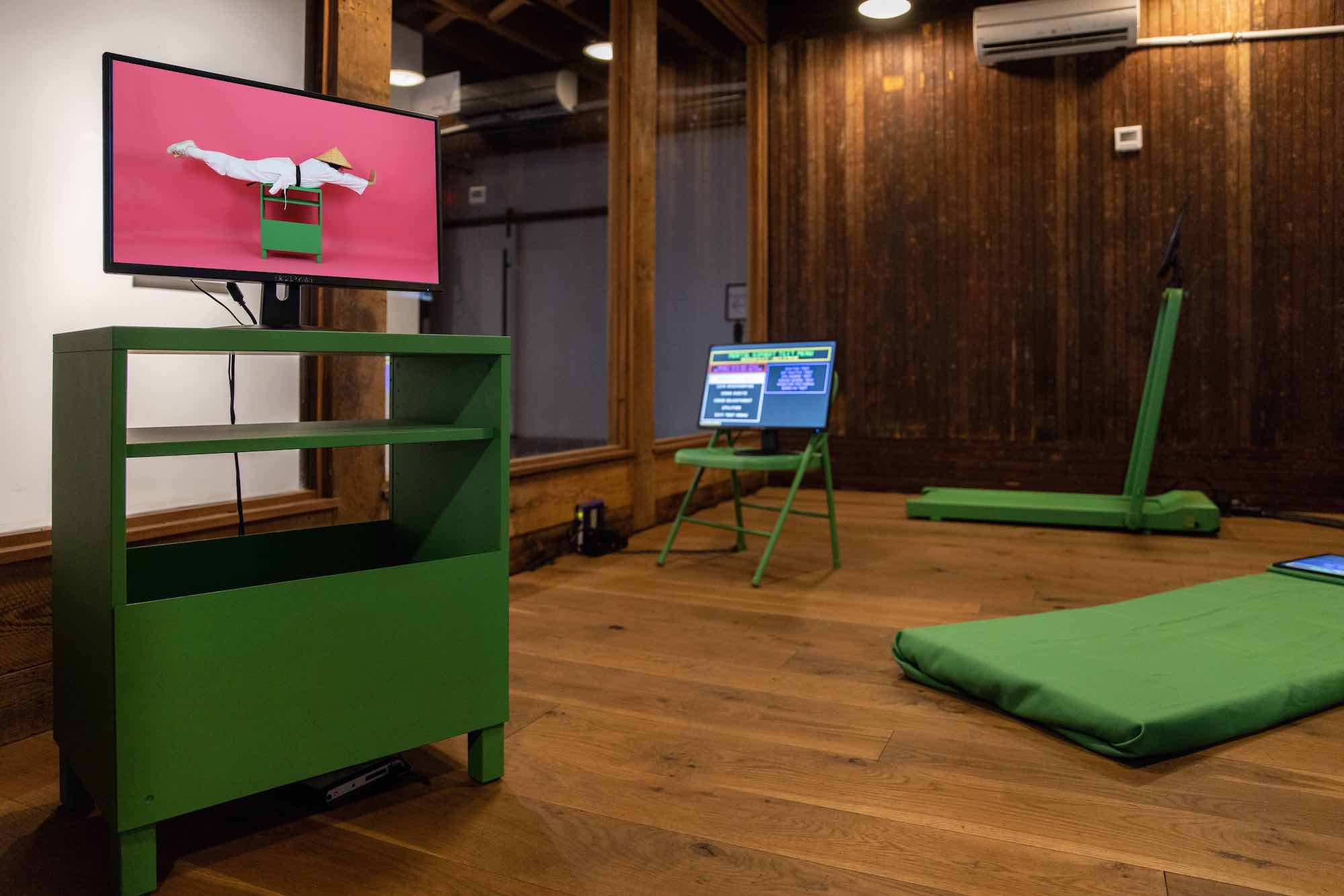 A room with chroma-key green furniture, each with a video monitor atop. The monitor in the foreground shows an image of a pink background with the artist laying belly down, outstretched on a green table wearing a white outfit and an Asian conical hat