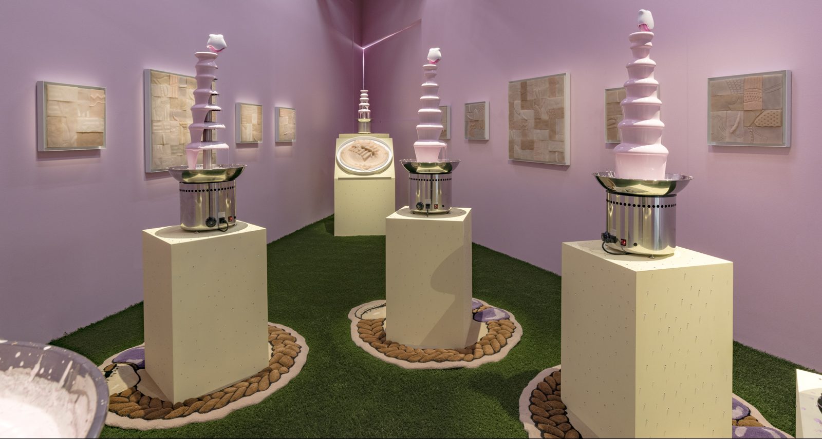 A light pink painted triangular room with green wall to wall astroturf flooring. From this perspective you can see three 6- tiered chocolate fondue fountains, but instead of chocolate they are filled with Lusters Pink Oil Moisturizer Lotion. Topping each of the fountains is a Sqweel 2 oral sex toy, the kind that is white with little pink silicone 