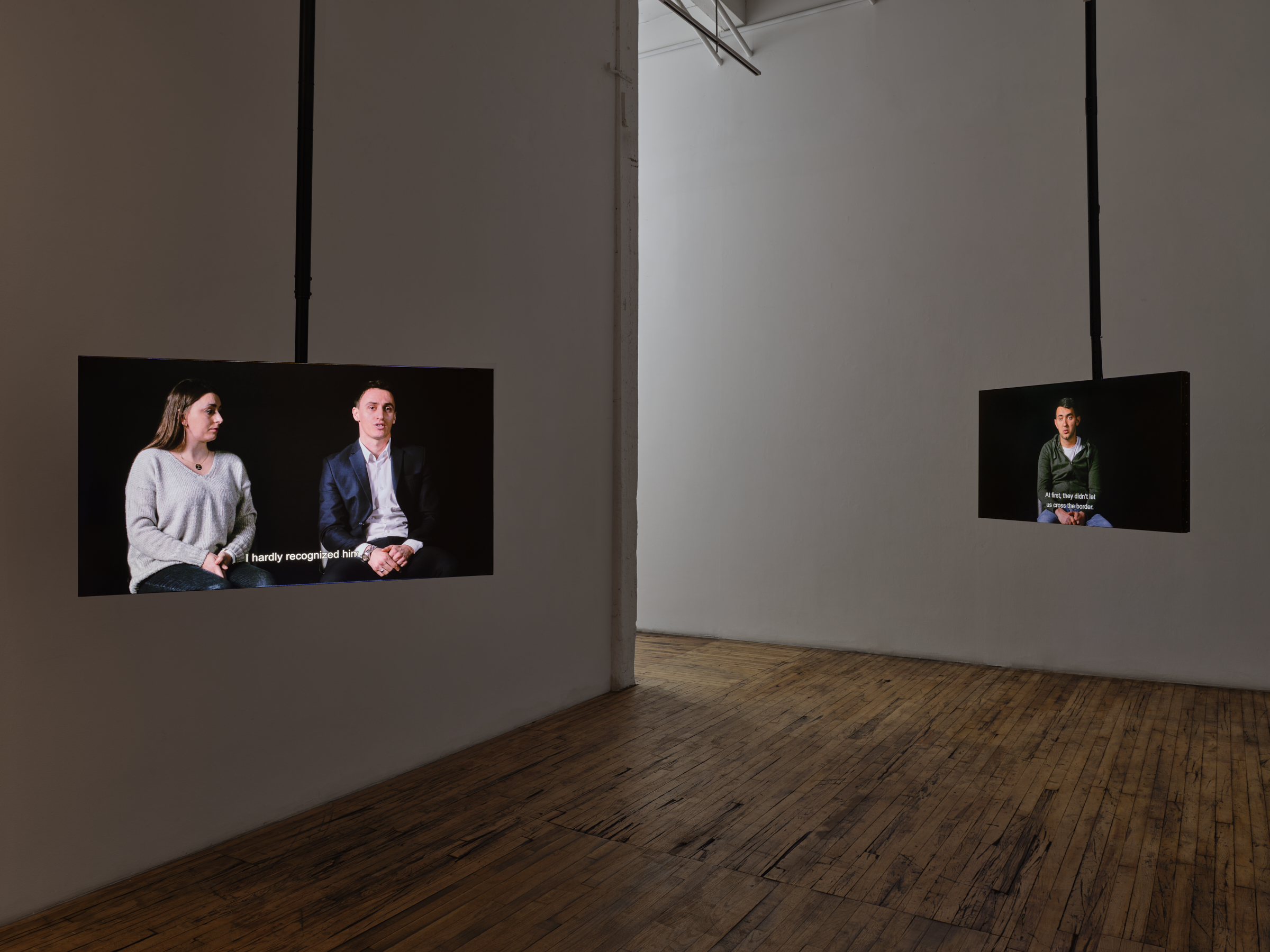 In a white room with wooden floors, two tv screens are hanging. The left screen shows two Kosovars talking to the camera, with the caption reading 