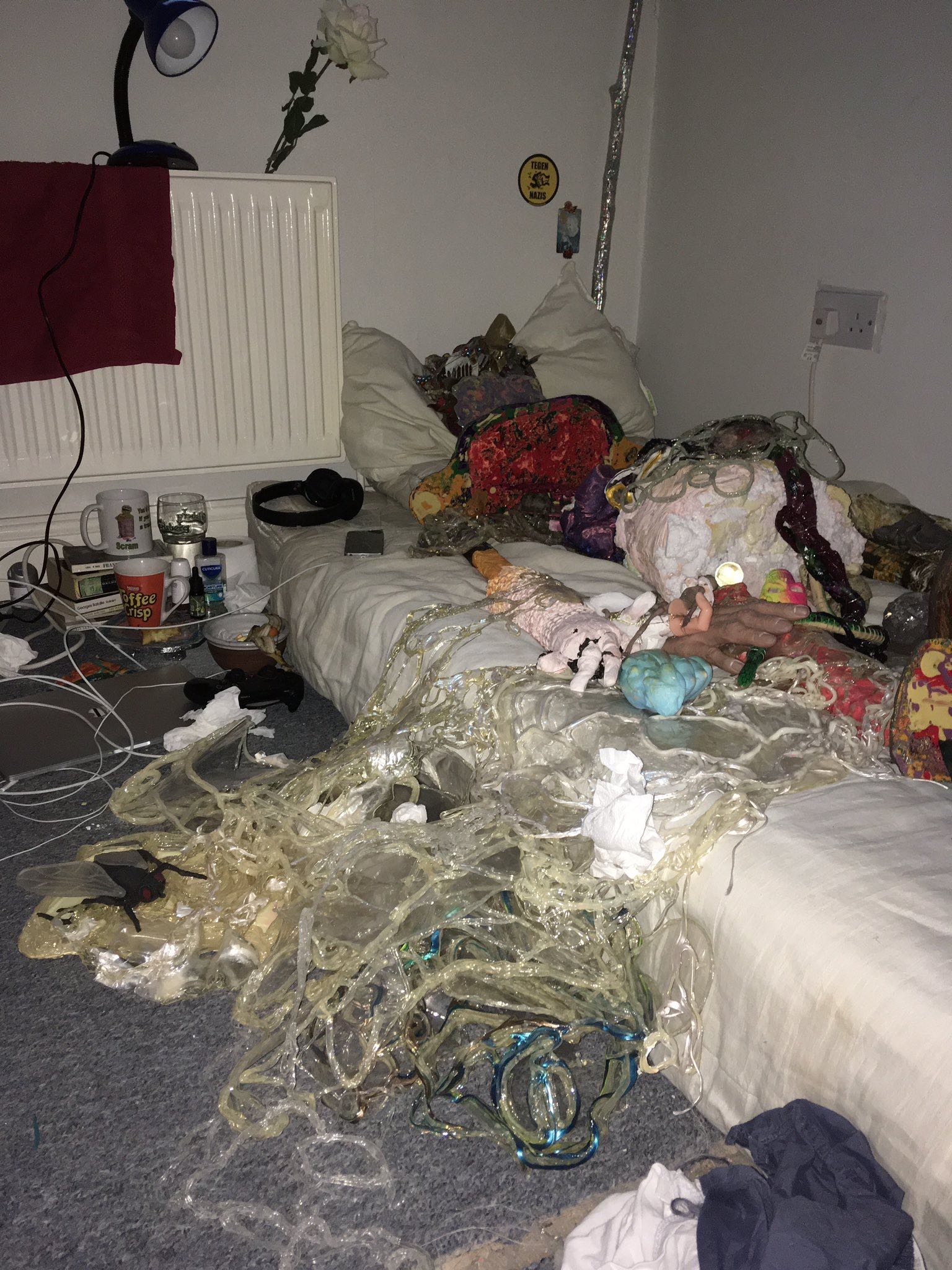 A photo taken with the flash on of the corner of a cluttered and messy bedroom. The mattress on the floor has no sheets and a figure lays on it made from a combination of smaller colourful sculptural objects and materials