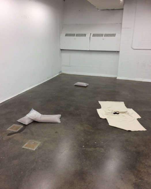 A gallery with stone floors and white walls. On the floor are various works: at the back and left foreground are salmon coloured pouches made of a cloudy plastic. In the right foreground is a cream-coloured t-shirt, cut at the side seams and laid flat.