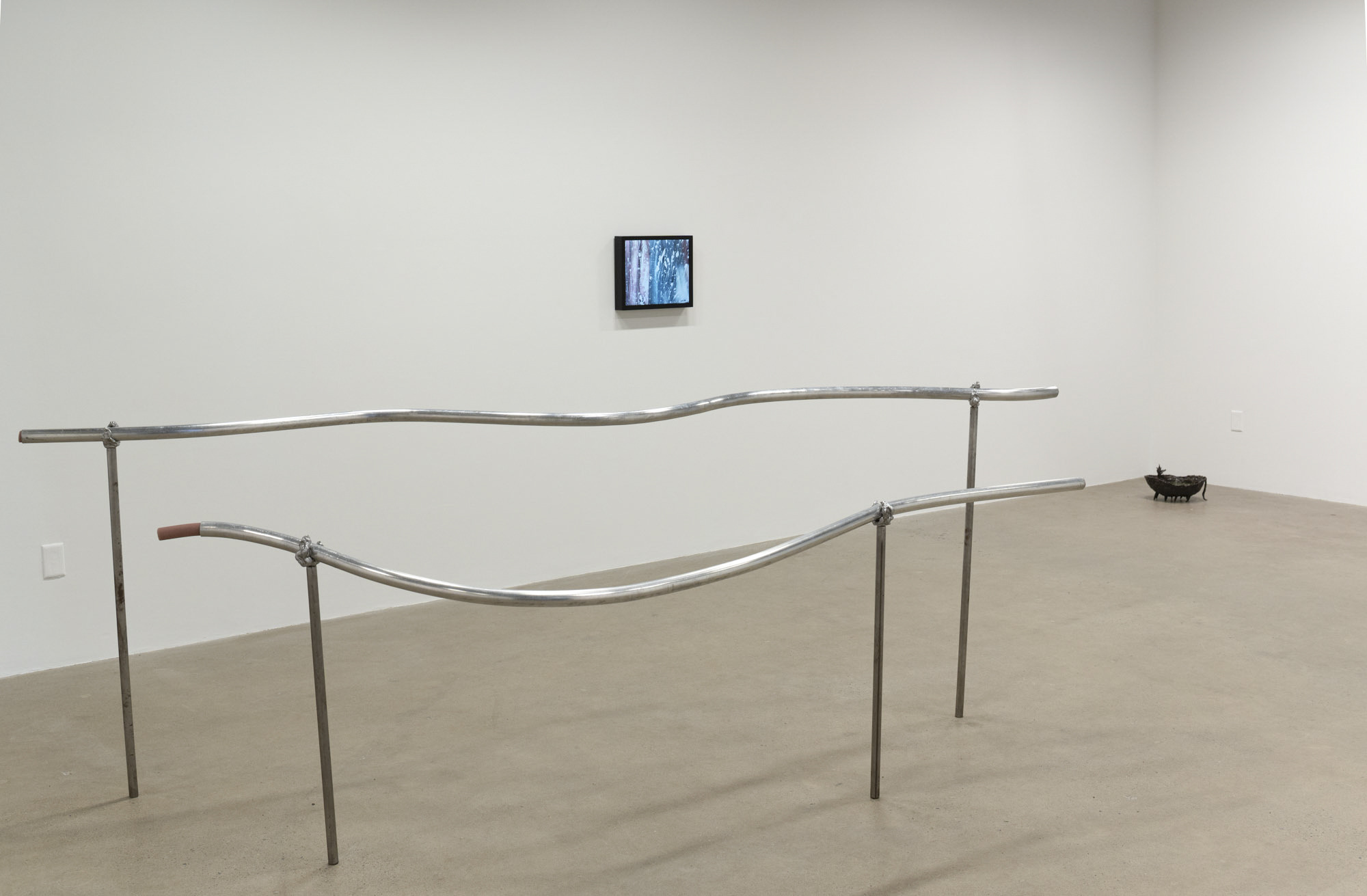 A gallery with concrete floors and white walls. A metal handrail sculpture is in the foreground. A monitor on the wall behind it shows a video work and a fountain sculpture in the far corner.