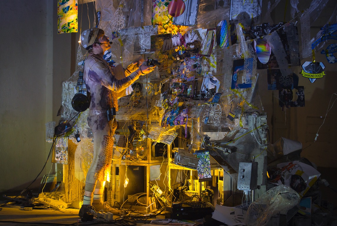 A man wearing a stained space suit gestures toward a large surface made up of keyboards, collaged images all covered in some kind of translucent slimy web