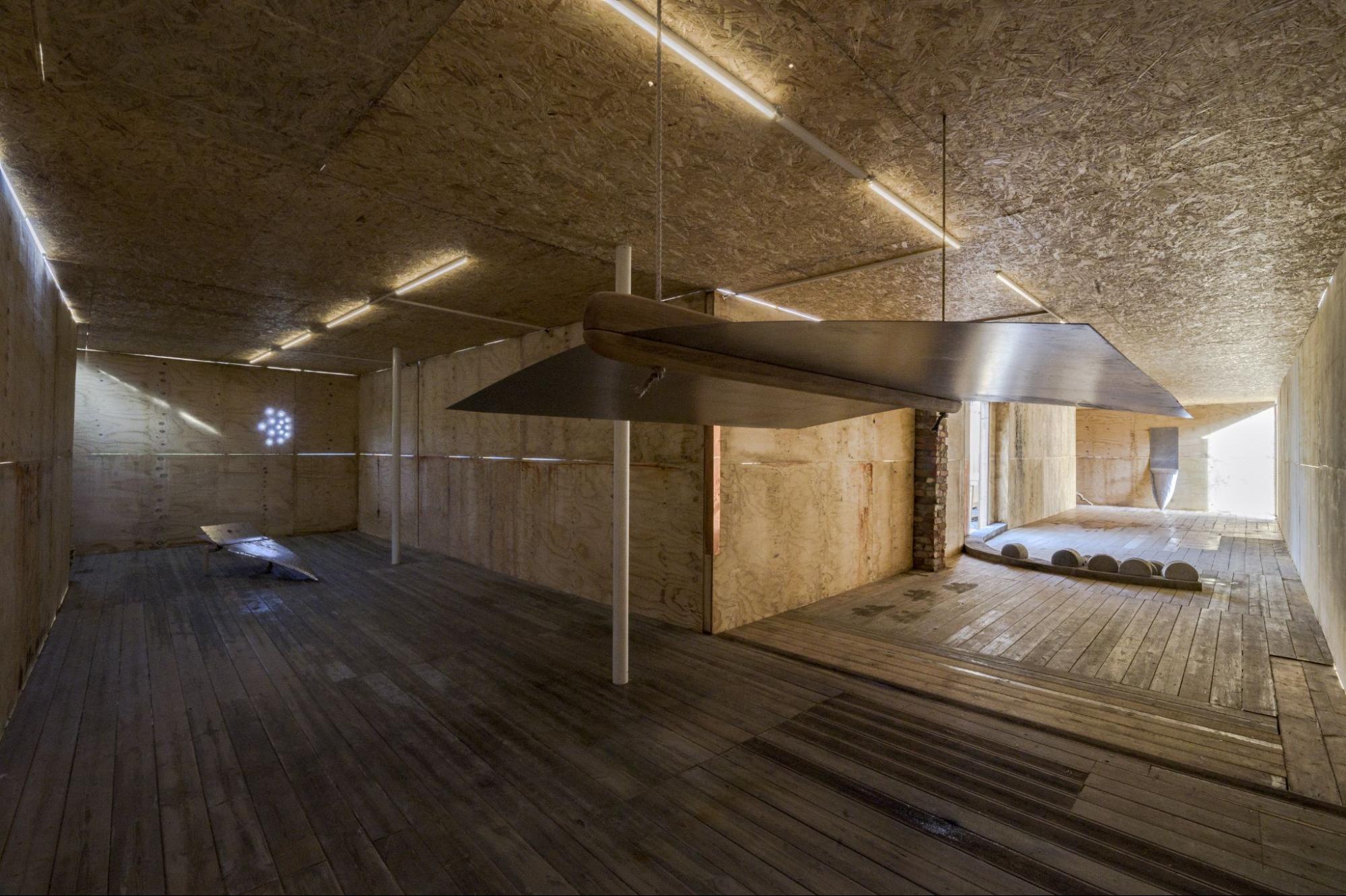 The image shows a room with fluorescent lighting, wooden floors, and plywood ceilings and walls. There are five sculptures exhibited in the space, fabricated with metal & brass, bricks, concrete, and ceramics.