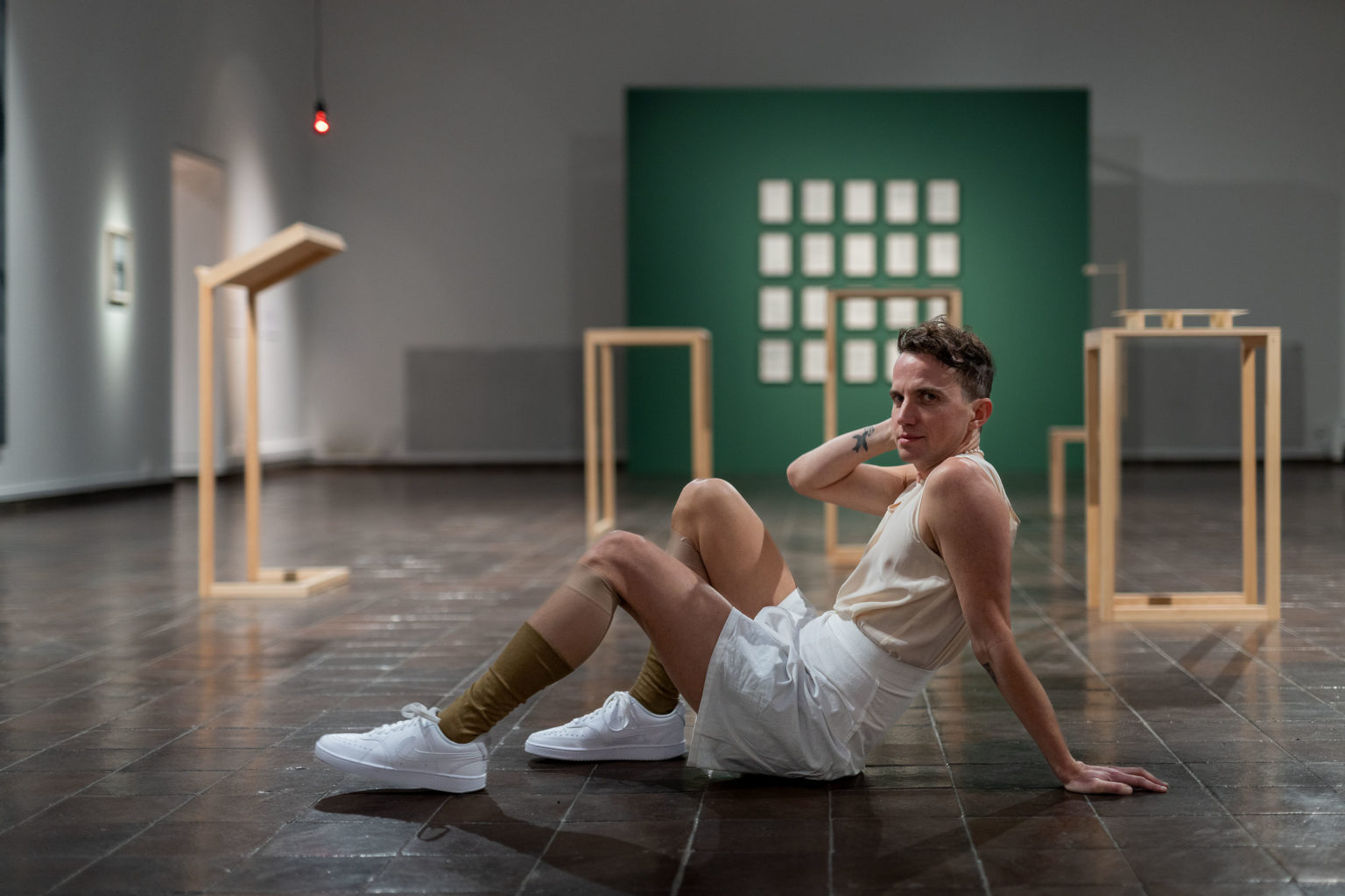 The image shows a young white man with curly light brown hair sitting on the floor of an art gallery in a reclining pose, looking into the camera. He has a tattoo on his inner right forearm and is wearing a light tan tank top, loosely fitting white shorts, high tan socks and white tennis shoes. Behind him we see minimal wooden plinths that display documents and an out of focus green wall with a grid of white papers on it.