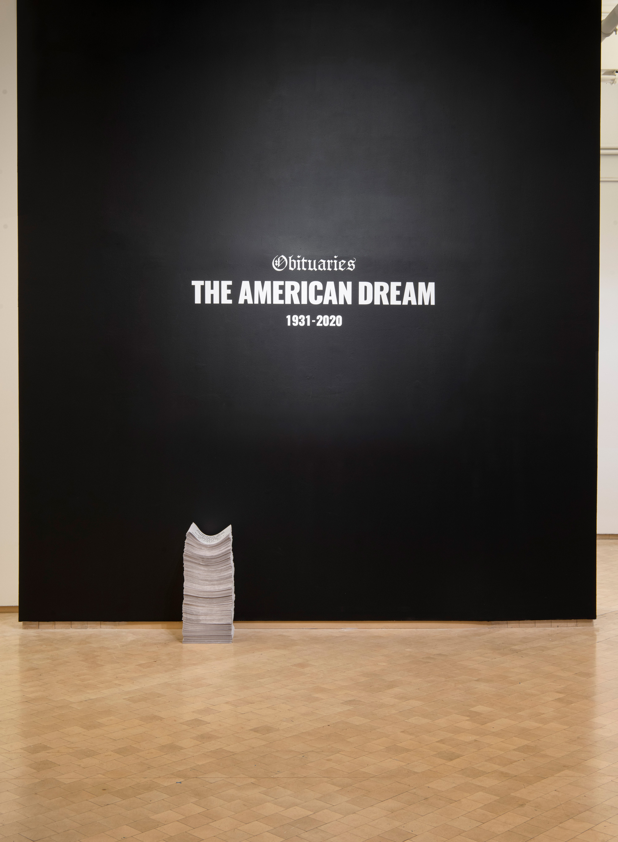 Installation of black wall with titled of the piece Obituaries of the American Dream along with newspaper print to take away