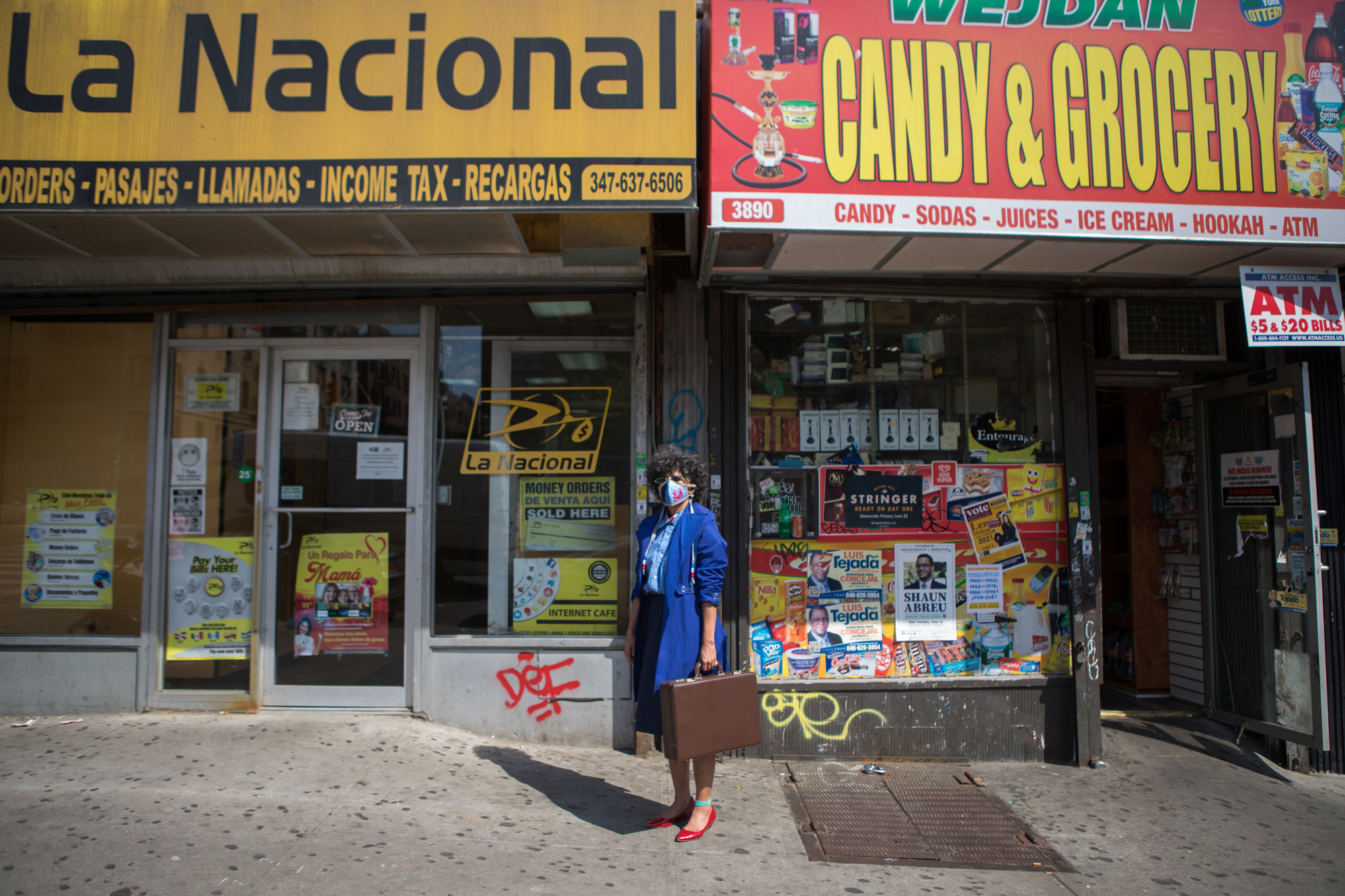 A photograph of a light-skinned Black woman with short, curly brown hair standing facing the camera. She is on a city sidewalk in between a brightly colored bodega and a check-cashing store. The woman is wearing a mid-length skirt, a button-down shirt, and a trench coat, all in varying shades of blue; red shoes; a red and blue face mask; and small, oval-shaped sunglasses. She carries a brown briefcase in her left hand.