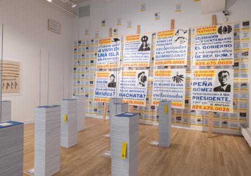 Installation view of exhibition. You can see a series of 20 books 5 feet tall and in the back a series of 8 posters on rice sack with blue, red, and yellow stamp letters.