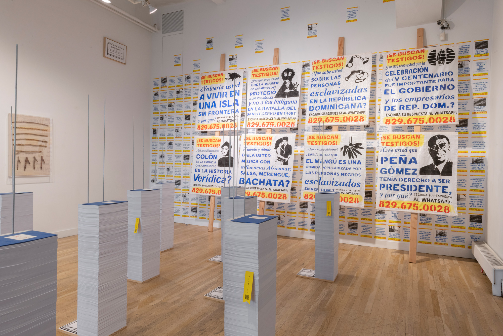 Installation view of exhibition. You can see a series of 20 books 5 feet tall and in the back a series of 8 posters on rice sack with blue, red, and yellow stamp letters.