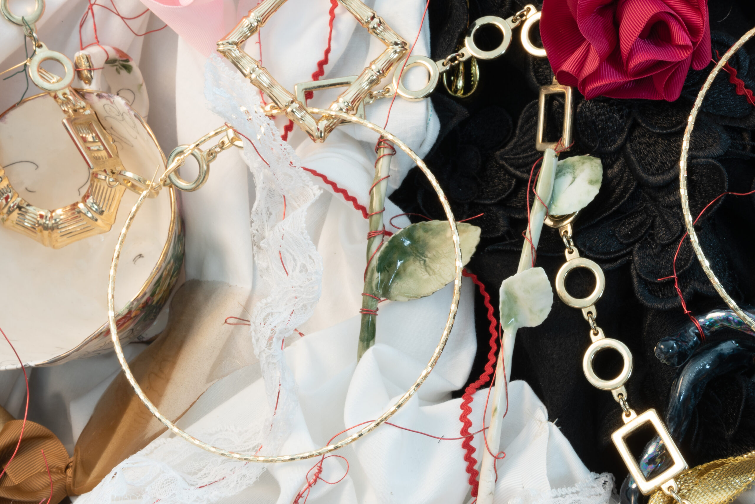 Adornments is the central work in the exhibition. It is a maximalist still life table installation consisting of an assortment of found objects and ceramic sculptures. Embroidered and manipulated fabric provide another textural layer. A detail photograph of the installation shows bright gold hoop earrings and chains laying on top of black and white fabric and lace. Strands of red thread are loosely wrapped around two green ceramic flower stems that are also intermingled with the jewelry; the red string continues throughout the image drawing our attention also to her ceramic teacup decorated with with the artist’s signature curly lines and to a part of her sculpture of a lock of curly hair. The work brings together Norwood's conceptual interest in the presence of Black bodies and labour in the histories of the decorative arts and in the intimate and public spaces that we take for granted.