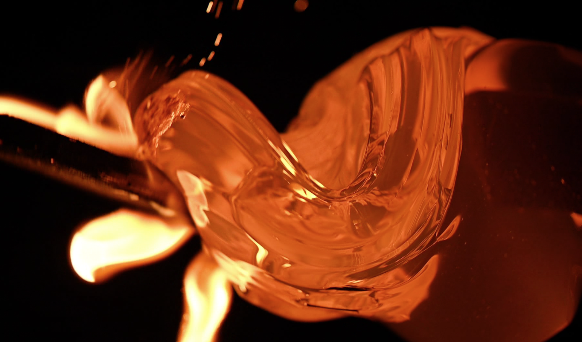 Ecstasy is a slow-motion video of molten glass. In the scene, the background is dark while the glass is illuminating the surrounding with heat. The molten glass on the end of a rod is turning slowly. The speed of the turning is not fast enough to keep the glass from falling. A wooden paddle comes into the frame from the left, slapping and pushing the glass from dropping. When the glass and wood meet, the heat burns the wood thus creating sparks. Sometimes the wooden pat stays longer on the glass, gently lifting the weight of the glass. As the two materials touch, the heat begins to light up the wooden paddle, creating a bigger flame than the sparks. The video is accompanied by the background sound of white waves crashing on shores.