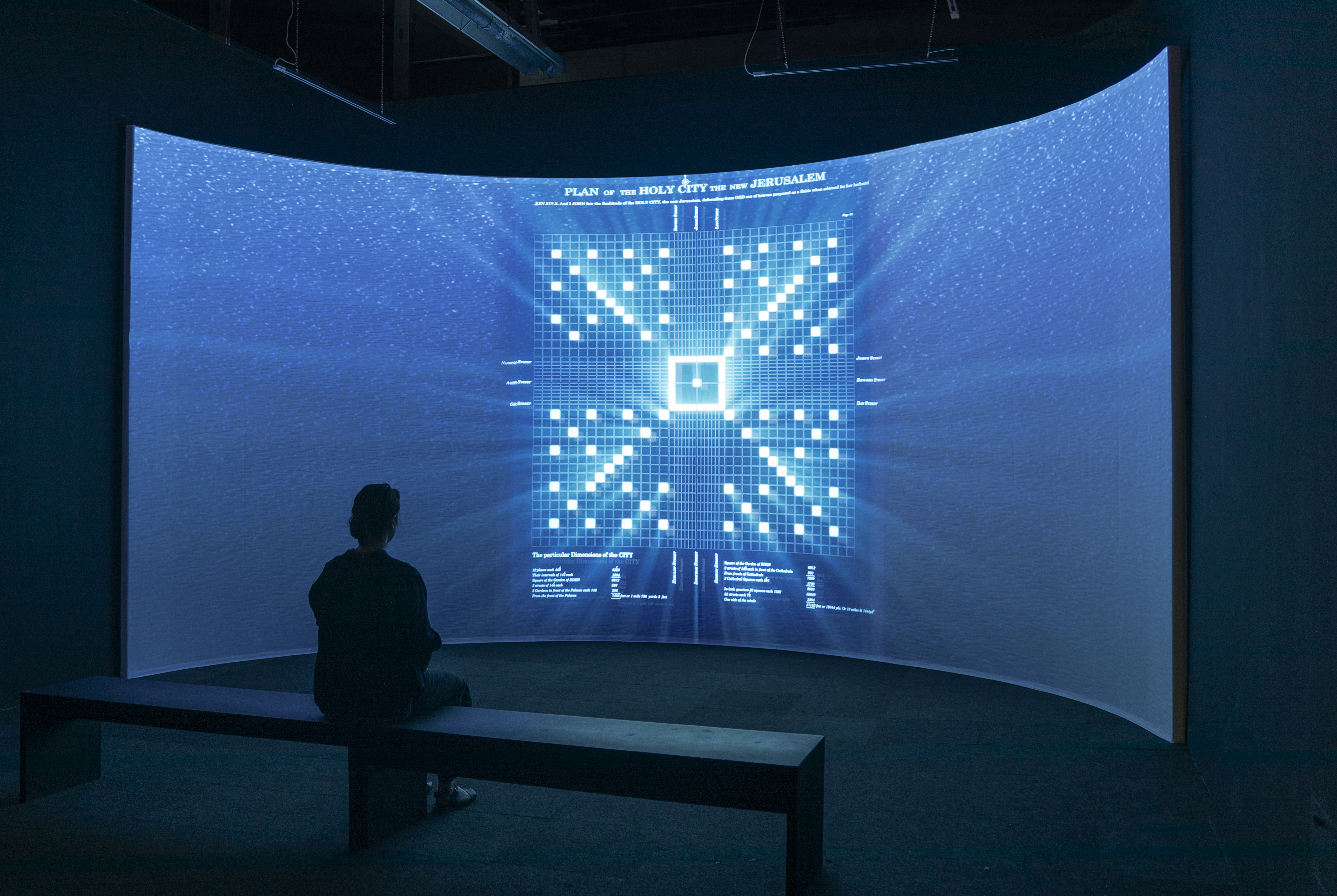Installation shot of a darkened space with a 180-degree curved screen with a projected image from Un-charting. A viewer sits on a bench in front of the screen with an image of a glowing whitye map against a blue water-like background depicting a 19th century map of Jerusalem by the self proclaimed British Prophet Richard Brothers.