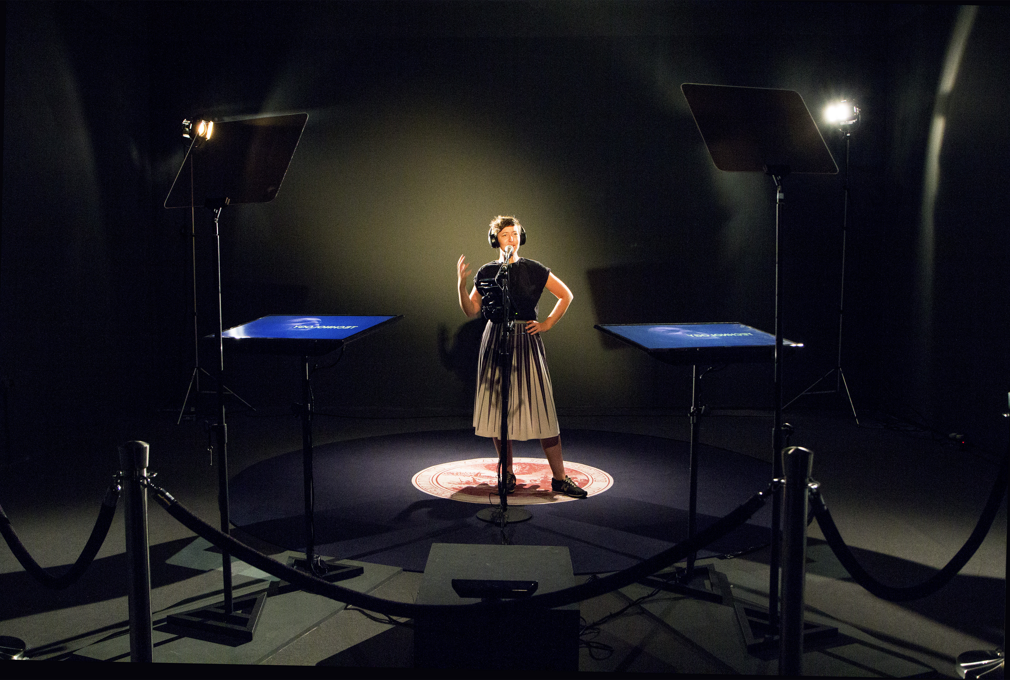 A woman standing in a dark room atop a regal rug imprinted with the rejected seal for the United States. In front of her are two teleprompters as she appears mid-speech.