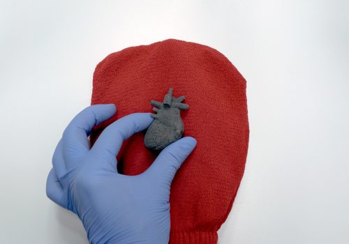 A charcoal bath bomb shaped like an anatomical heart is gently held by a gloved hand.