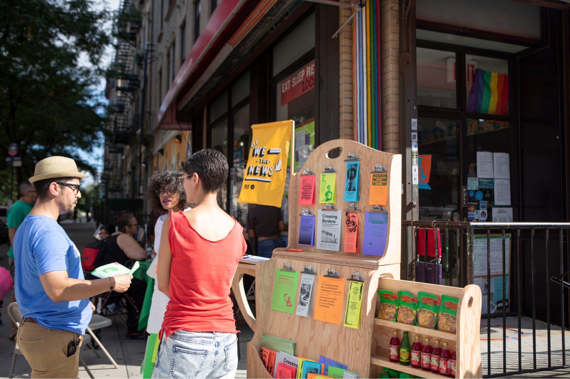 A wooden newsstand festooned with brightly colored paper booklets and containing shelves of hot sauce and snacks stand on a New York City sidewalk in the sunshine. To the left of it a White woman and Latinx man stand with their backs to the camera facing the artist, an Afrolatina woman, who is speaking to them.