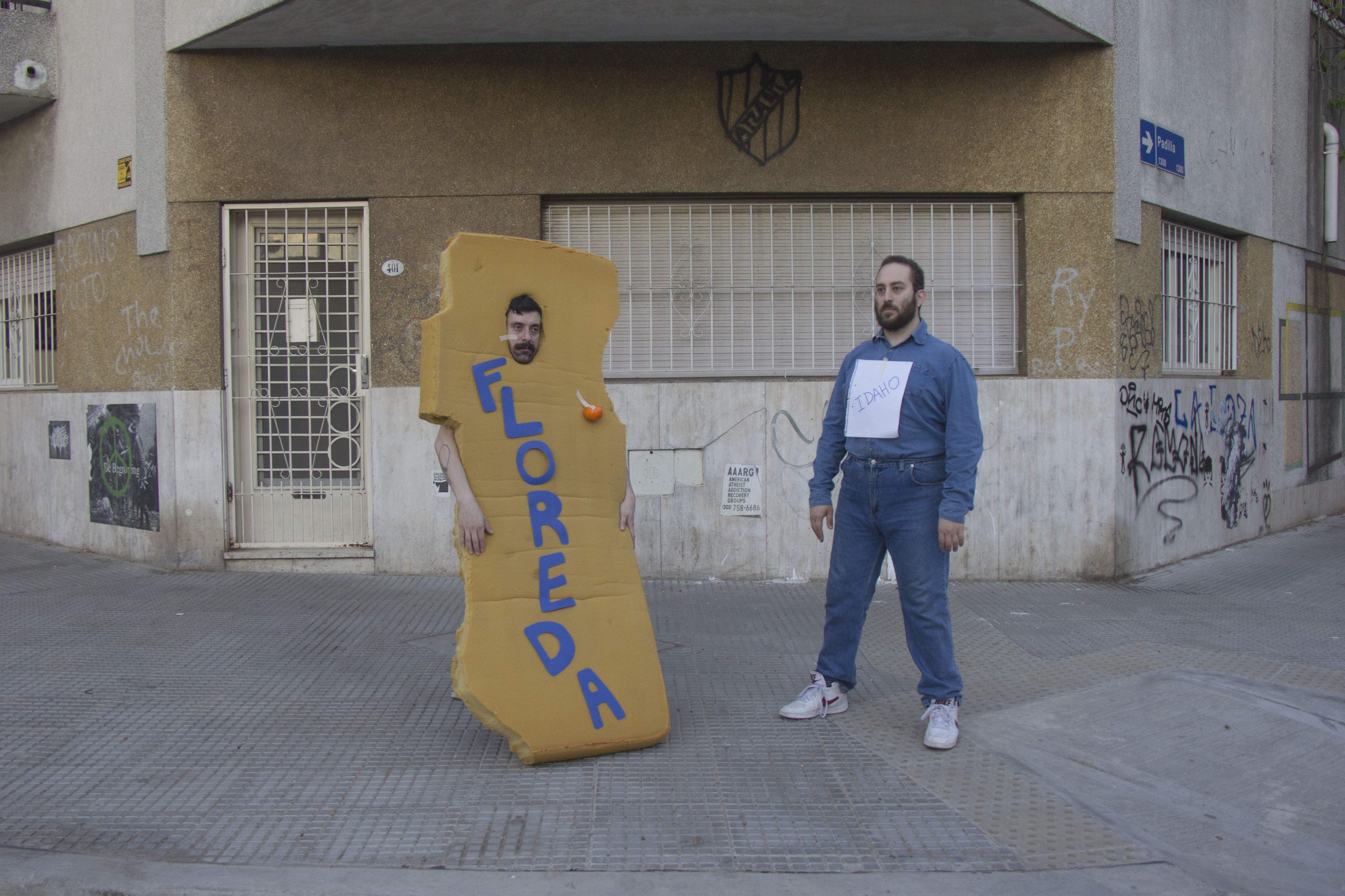 This is an exterior photo of a concrete building with metal bars over the windows and some graffiti. In the foreground there are two figures: one White man with a mustache wearing a large foam costume with the text “FLOREDA” spelled out on it in blue block letters. The other White man wears a beard, an all denim outfit, and has a white piece of paper on his shirt that says “IDAHO.”