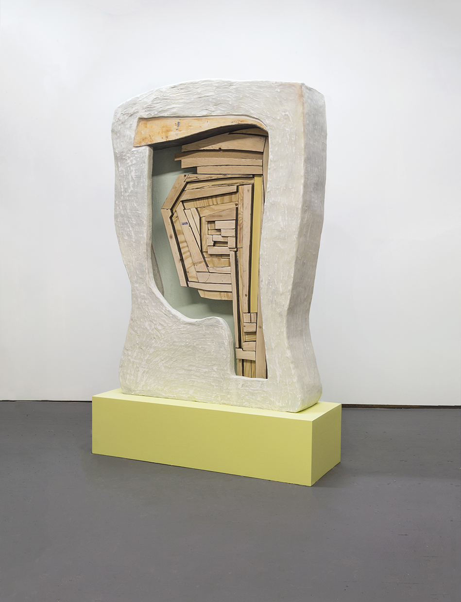 A life-size faded, grey, stone-textured vertical stele form sits atop a subdued lime green, smooth-textured rectangular pedestal which sits flat on the floor. The stele form is subtly curved in the shape of a gravestone. There is an organic, but rectangular shaped cutout in the center of the stele form, revealing its interior as well as a mosaic of wood fragments that are framed within the shallow interior of the cutout. The wood fragments are plywood, pine, and hardboard. Some of them are treated with a green or black paint wash. The wood mosaic forms taper off inside the form and consolidate toward the base.