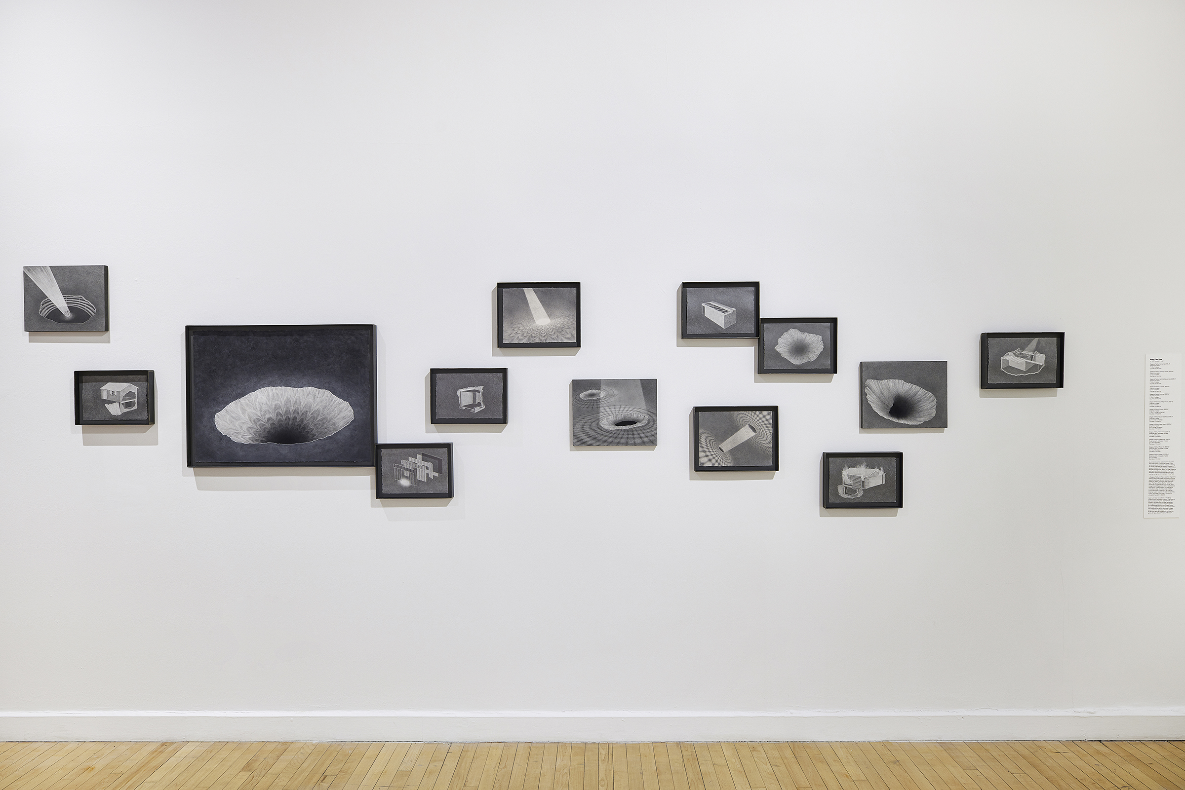 Installation shot of “Stages of Fallout” showing a constellation of graphite drawings hanging against a white wall at various heights. Each drawing is framed in a grey steel frame.