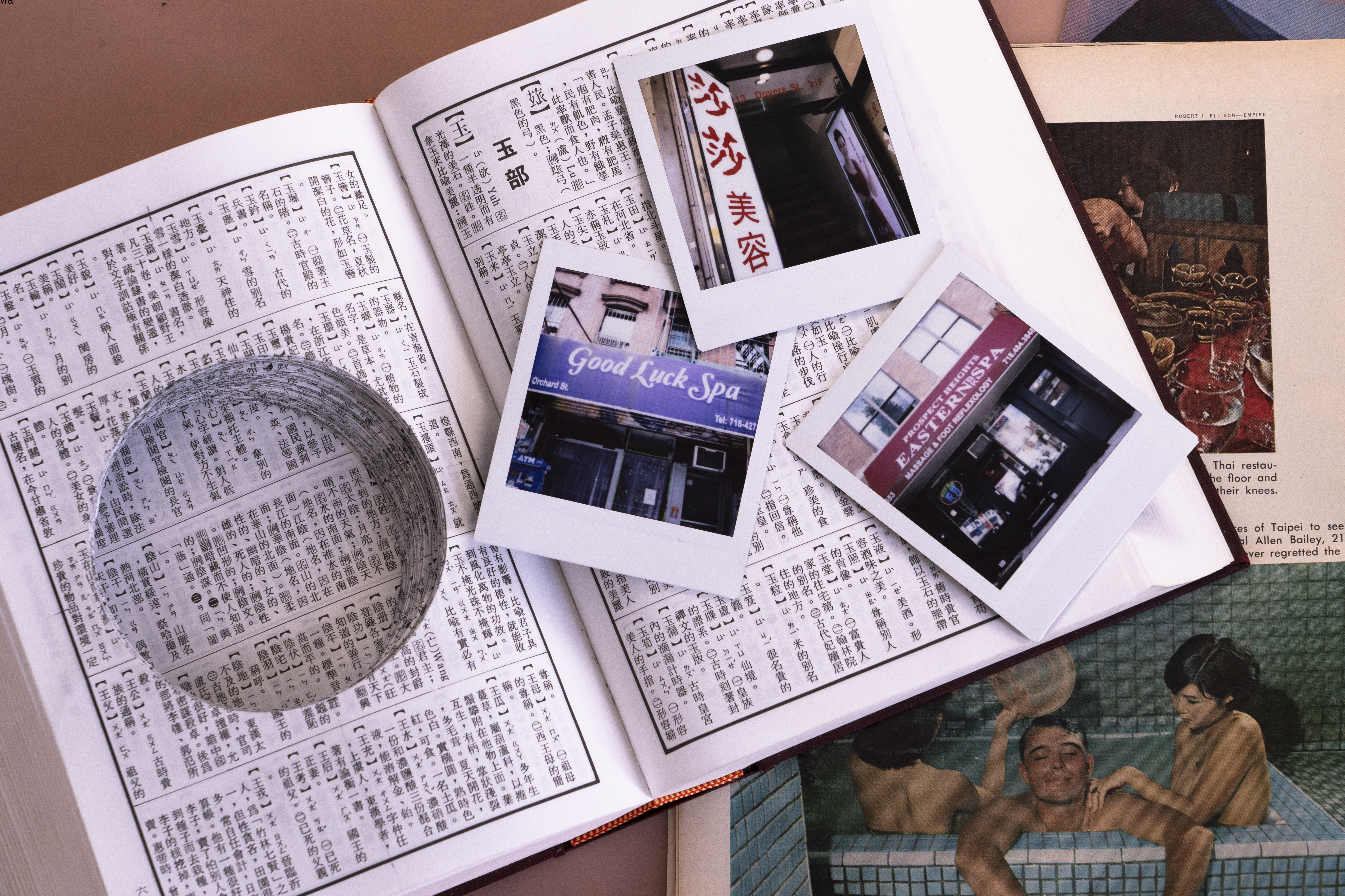 Overhead photo of an open Taiwanese dictionary. The pages on the left side of the book have been neatly cut with circular holes, creating a precise empty cylinder, about one inch in depth. Three polaroids are laid on top of the right side of the book, each shows the sign for an Asian massage parlor: “Good Luck Spa”, “Prospect Heights EASTERN SPA Massage and Foot Reflexology” and a sign in Chinese characters. Below the dictionary there is an open book that shows a Time magazine photo of a White man in a Japanese style tiled bath with young Asian spa workers massaging and bathing him.