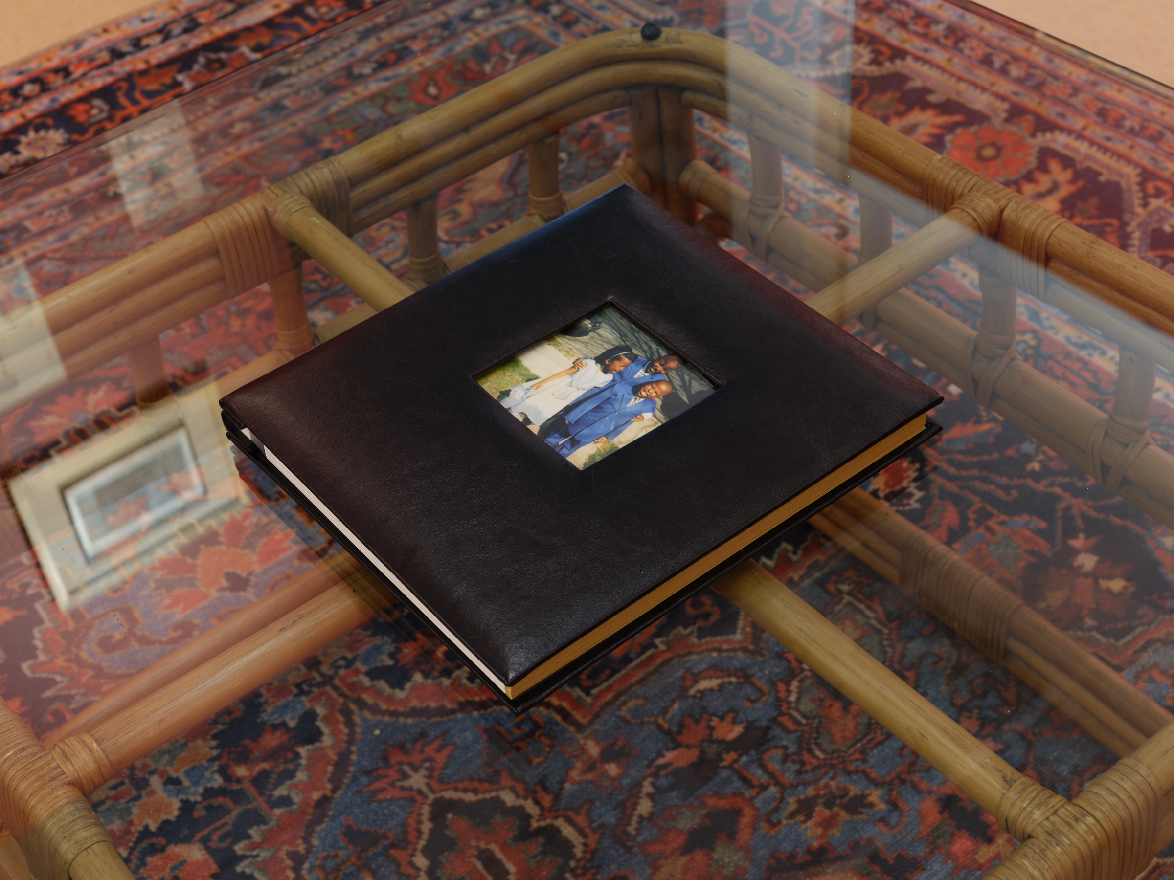 A black leather bound photo album with a picture of three young Black children dressed for church rests on a glass top coffee table. The patterned red, blue and black carpet below is visible and some reflections of framed artwork from the show can also be seen.