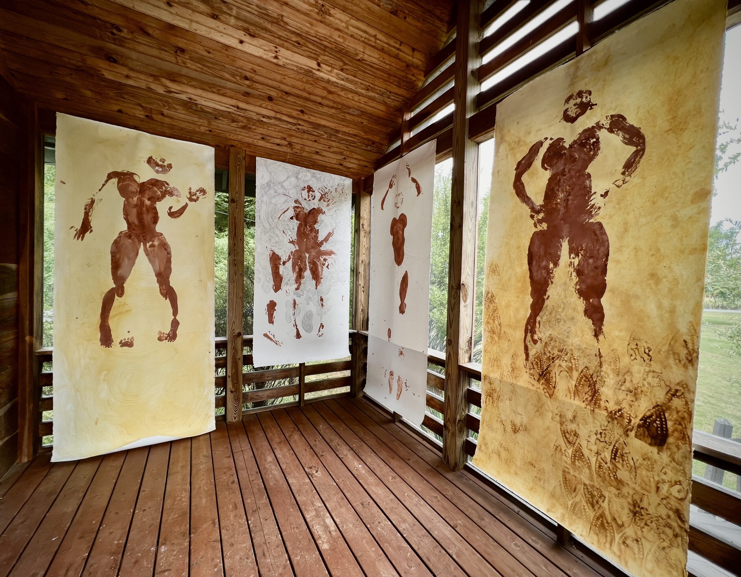 Four large sheets of paper are hanging in a wooden screened in porch. Each has an impression of the artist’s body in dark red paint in the center of the paper. The outer two works have a turmeric-colored background and one is marked with burns from a clothes iron. The two inner works are on a white background and one has pen drawings in the negative space.