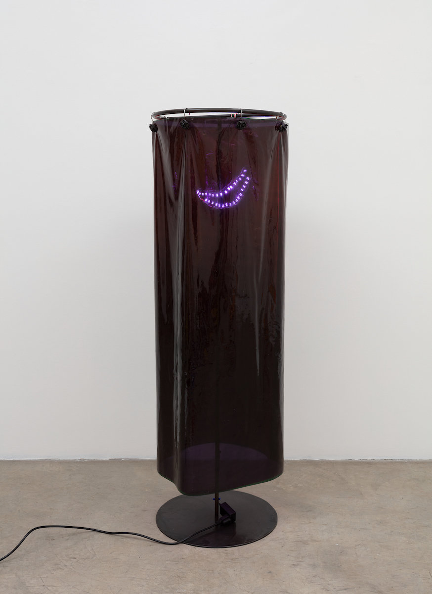 A shiny, semi transparent black plastic cylinder made with vinyl attached to a black metal stand sits on a smooth concrete floor. A wire leads into the cylinder from the bottom left corner and powers a small string of purple LED lights that are suspended in the in ner top area of the cylinder.