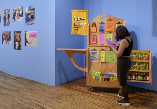 A woman wearing black clothing stands reading a booklet in front of a wooden newsstand cart that displays colorful publications. There is a yellow flag attached to the newsstand that says “WE THE NEWS.” The wall behind is a light shade of blue and to the left of the stand there are some photographs of people participating in the project mounted on the wall.