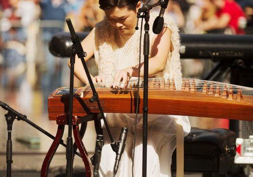 Clae Lu performing on the guzheng (Chinese zither) with a large audience at the Metropolitan Museum of Art.