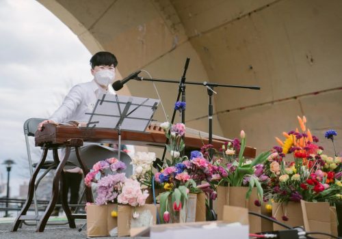 Clae Lu sitting behind their gu zheng in the amphitheater at East River Park. They are wearing a white mask, white shirt and light gray pants. In front of them are a variety of colorful flowers in brown bags