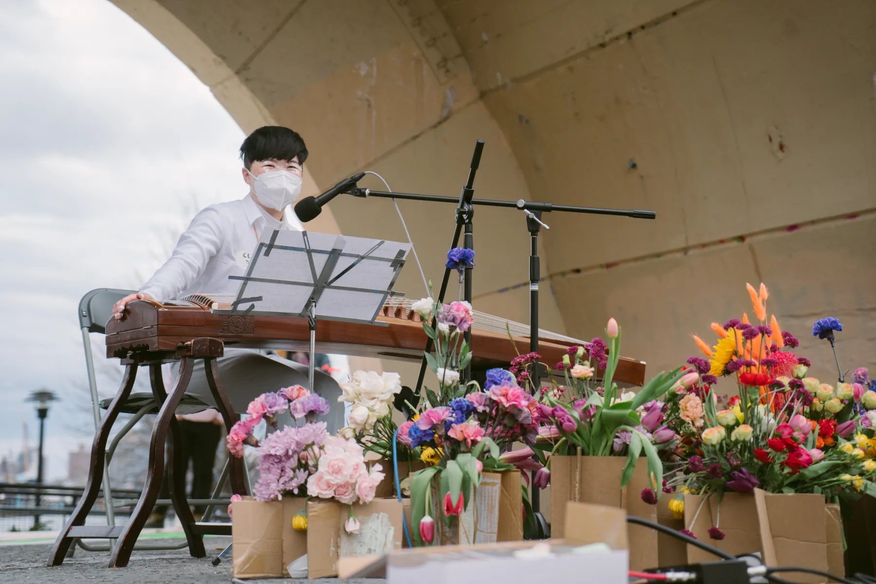 Clae Lu sitting behind their gu zheng in the amphitheater at East River Park. They are wearing a white mask, white shirt and light gray pants. In front of them are a variety of colorful flowers in brown bags