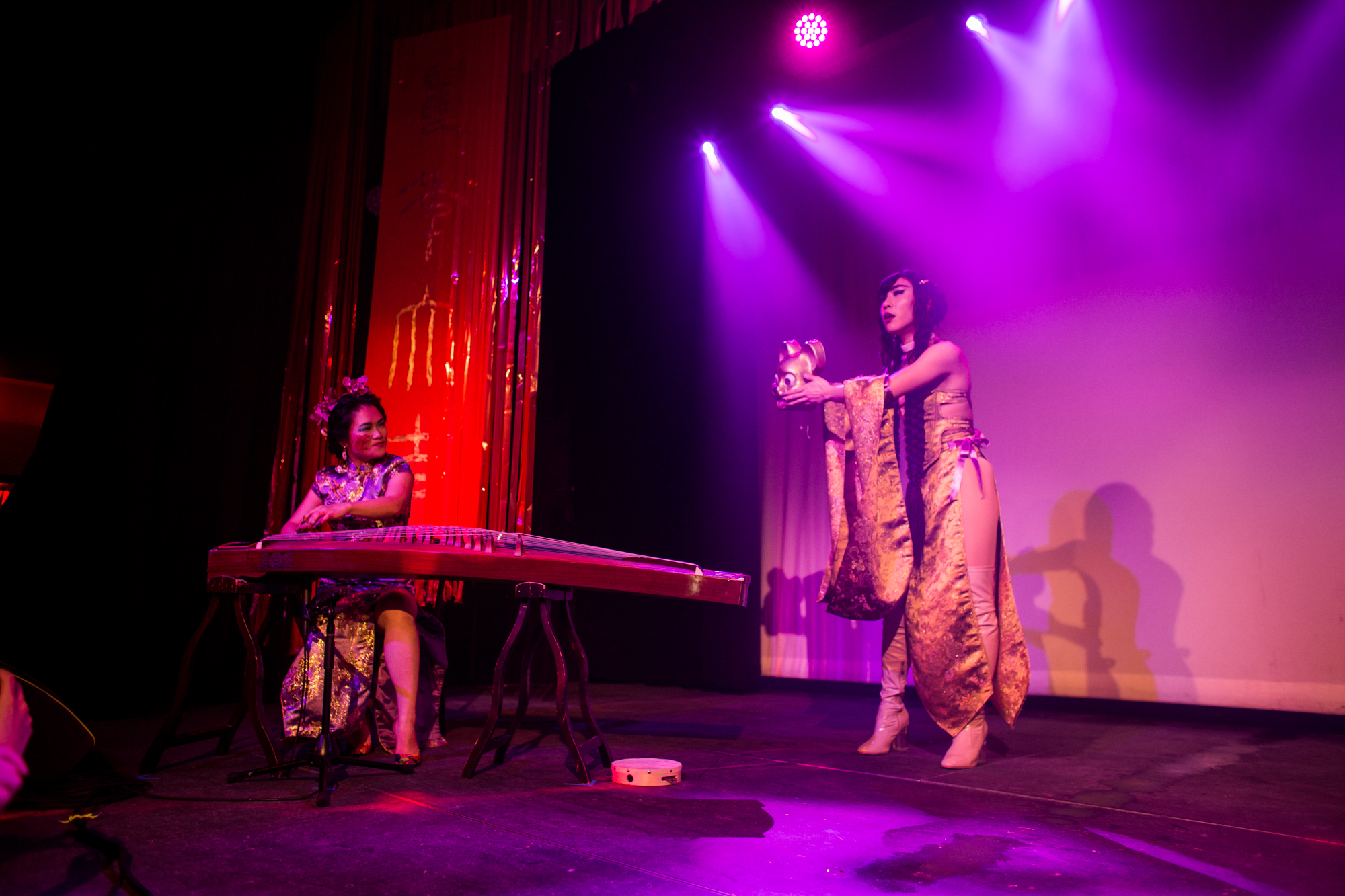 Clae and Dynasty performing on stage at Elsewhere in Brooklyn. They are lit by purple and red lights. Clae is in a purple qipao, performing on the guzheng looking at Dynasty who is holding a rat mask and in a gold costume.