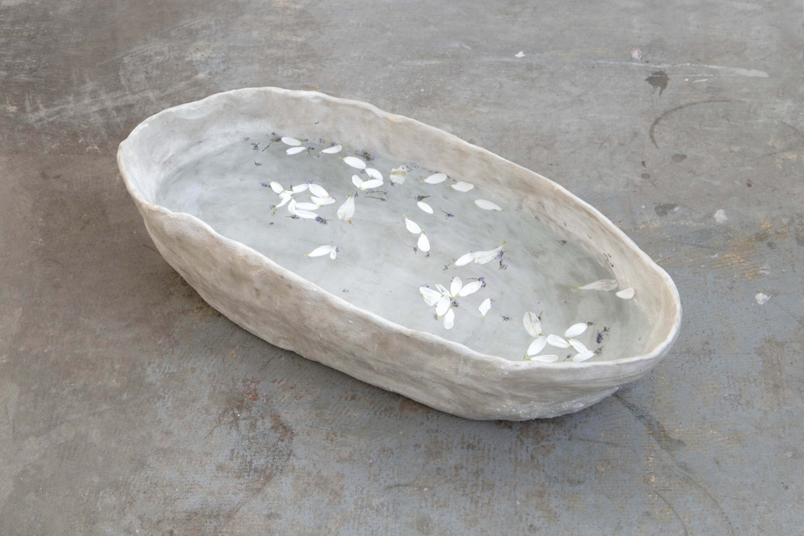 Ceramic bathtub displayed on the floor. The work has a textured grey glaze. The basin is filled with water and has white flowers, mint, and lavender floating inside.