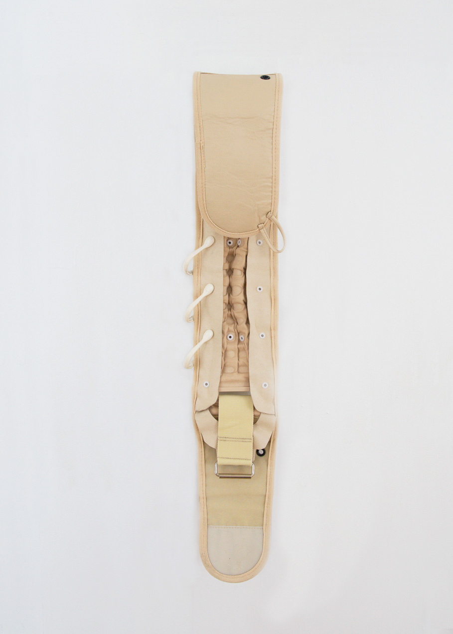 A long beige sculpture hanging on the wall. This work resembles a spinal cord with different textured fabrics of the same tone sewn together. On the side are three plastic rings.