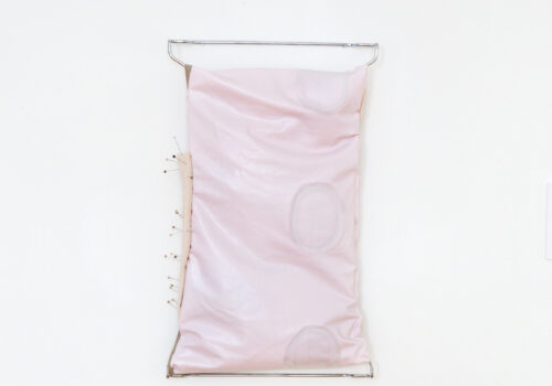 A pink air mattress sewn onto a metal rectangular structure. The left side of the work has a beige patch of fabric with numerous ball point sewing pins protruding from it. This piece is displayed on the wall.