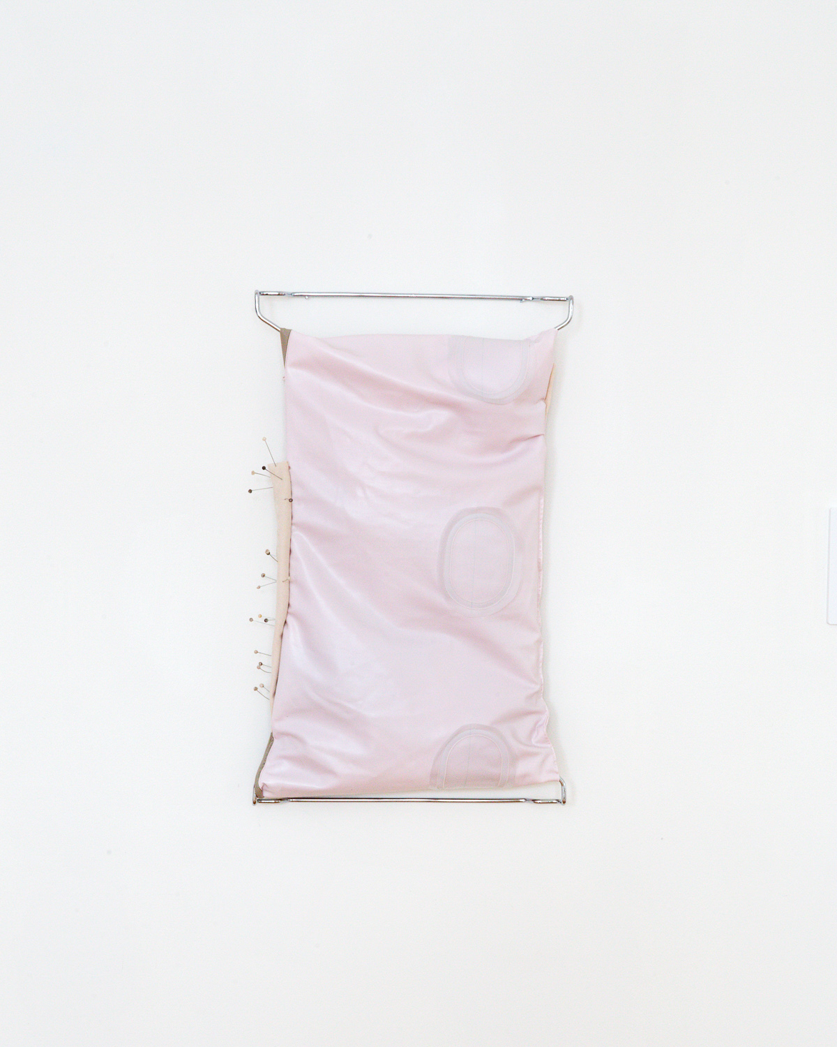 A pink air mattress sewn onto a metal rectangular structure. The left side of the work has a beige patch of fabric with numerous ball point sewing pins protruding from it. This piece is displayed on the wall.