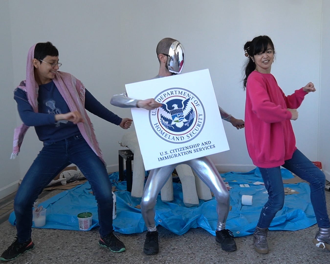 Three people dancing in sync. The middle is wearing a silver unitard, silver mask, and a USCIS logo.