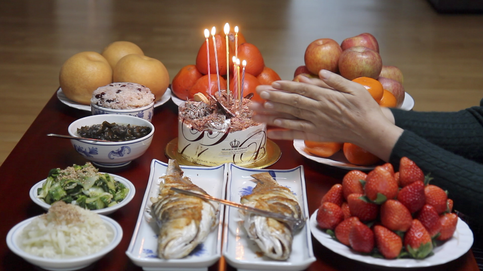 This is a photograph of a Korean dining table set with a small, fancy birthday cake with tall candles. Around the cake are plates piled high with strawberries, oranges, apples, persimmons and Korean pears. There are also two gilled fish and smaller bowls of rice, green vegetables, and black beans. From the right side of the frame, someone holds their hands in a gesture of prayer over the meal.