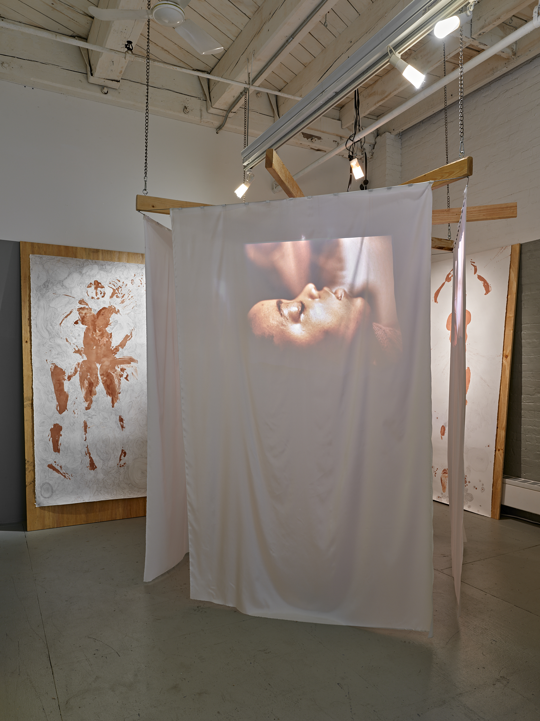 This is a view of the gallery installation which shows a video still that is rear projected onto a white sheet of the face of a young Black woman at rest with her eyes closed. The sheet hangs from a wooden structure suspended from the ceiling. Behind it there is a brown body print with line drawings around it on a large sheet of white paper mounted on a plywood sheet.