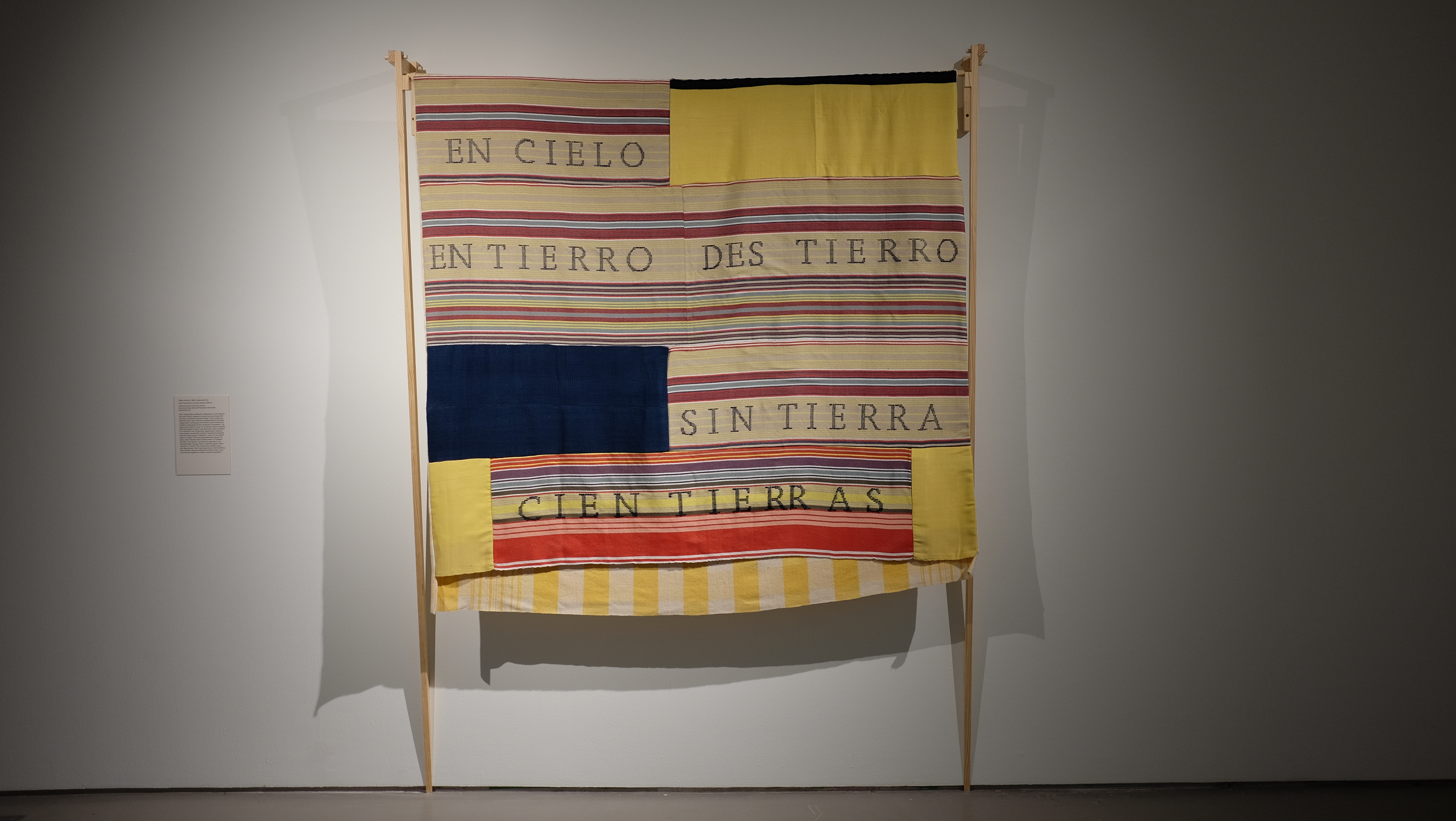 Cien Tierras features different configurations of the Spanish root word ‘tierra’ rendered in brown and navy thread with red, blue, and yellow horizontal stripes. The brocaded text features poetic uses of literary wordplay devices including malapropism, orthography and idioms to demonstrate the pliability of language and the complexity of translation. The upper panel reads ‘en cielo’ or ‘in the sky’, which is abutted with the phrases ‘en tierro’ and ‘des tierro’ which translates to ‘in the land’ and ‘of the land’ respectively. However, when spoken sounds like ‘entierro’ resembles the Spanish word for burial or grave. ‘Destierro’ means to be pushed away or exiled. The lower panel depicts the phrase ‘sin tierra’ which literally means without land or landless. The title, ‘cien tierras’, connotes the xpression ‘one hundred lands,’ though tierra could also signify earth, ground or world.