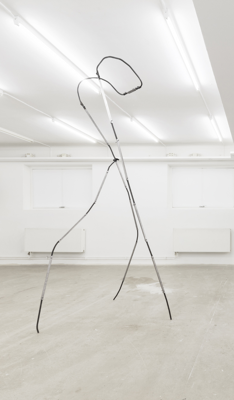Tall steel sculpture on three legs standing in a white room with concrete floors