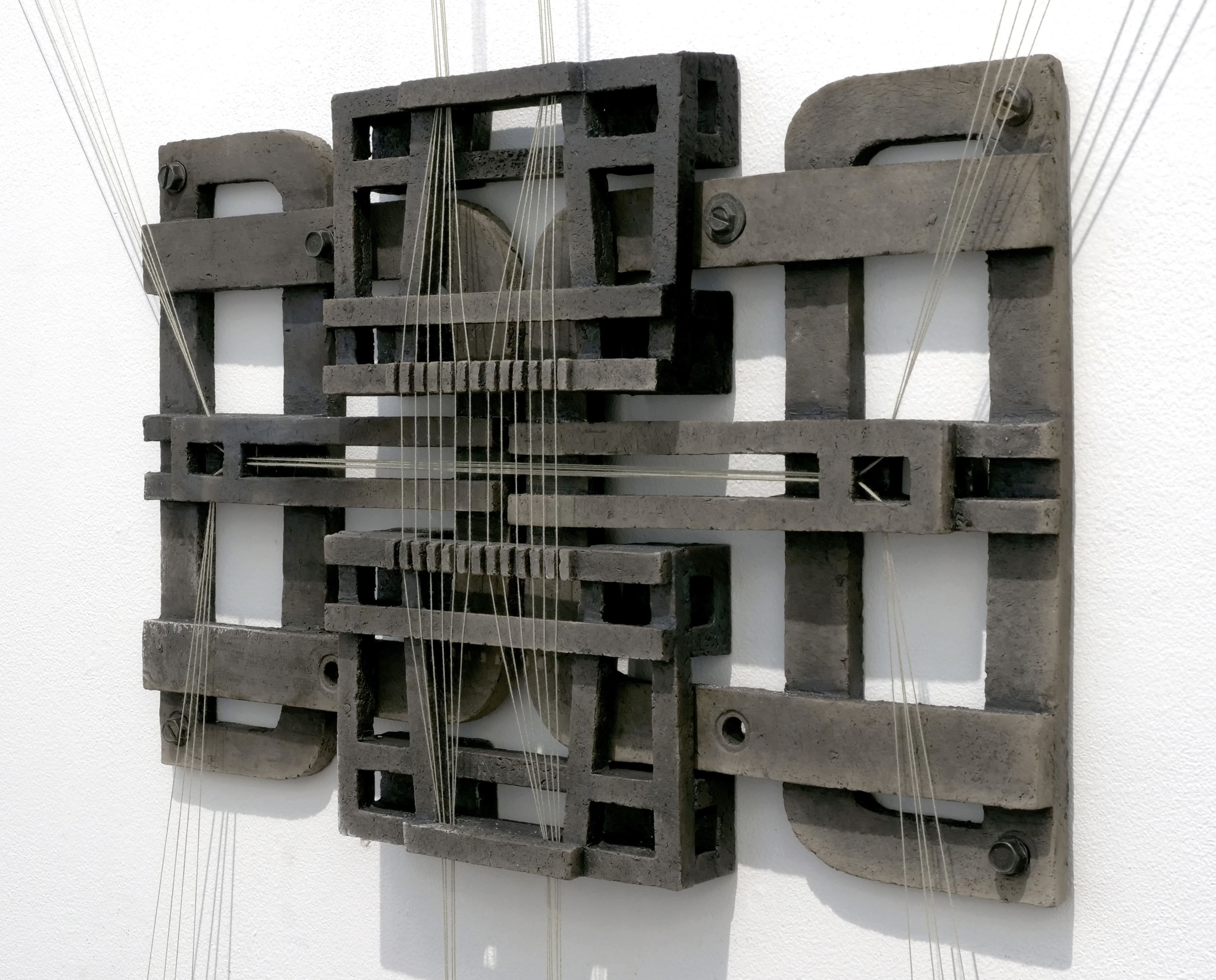 This is a wall mounted sculpture made of dark gray matte fired ceramic. The pieces appear to be part of industrial machinery with bolts fastening them together and white strings running through them at precise intervals.