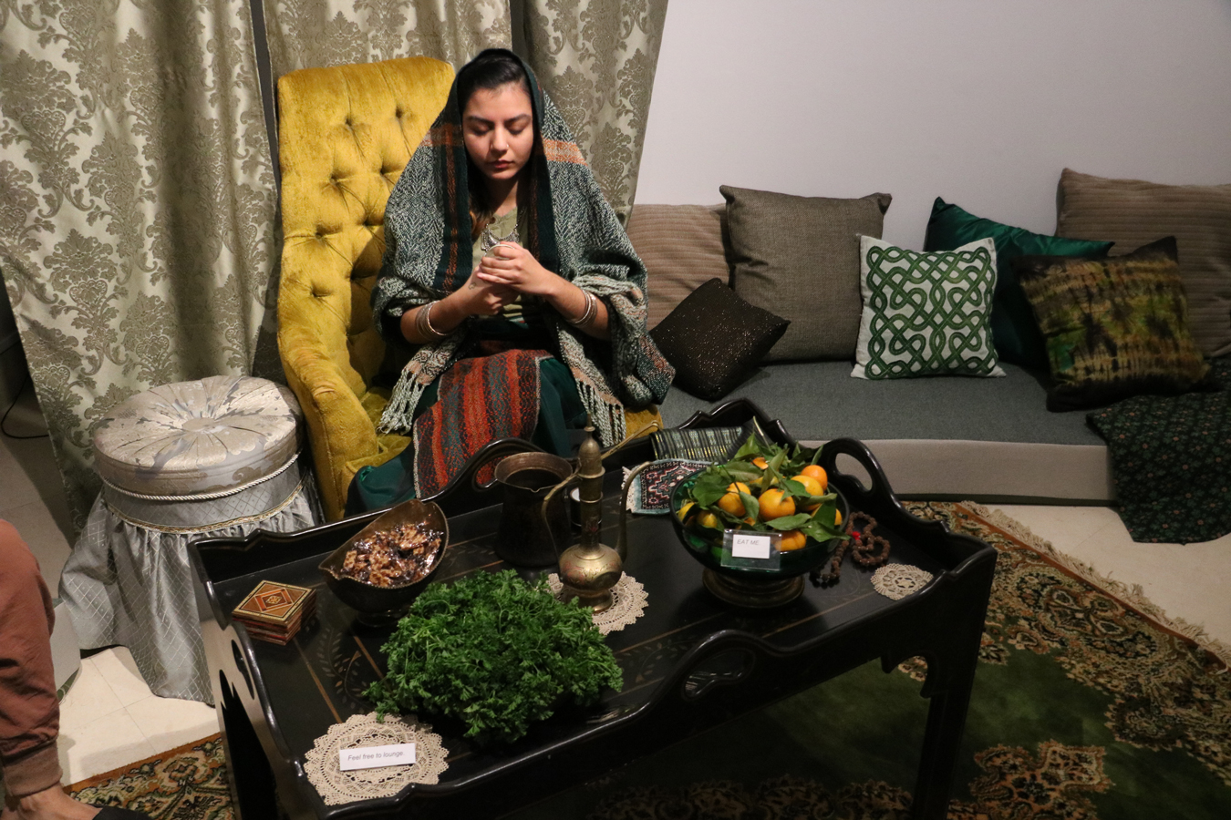 A figure dressed in various green handwoven and found fabrics, reminiscent of Southwest Asian traditional clothing peers into a demitasse coffee cup to ascertain the future. She sits within a living room installation in emerald and rust tones behind a table set with small oranges, parsley, sesame candies, hand-knotted doilies and decorative brass vessels.