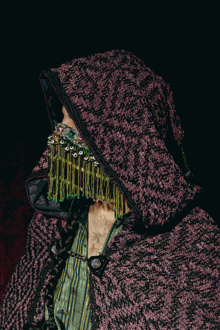 A cloaked figure is depicted torso-up with face obscured by a large draped hood and a face veil encrusted with beads, edged with evil eyes and beaded fringe in green and purple. The figure wears a handwoven cloak with a diamond weave pattern in a reddish-pruple and black textured patterning, with a bejeweled black clasp and a horizontal striped silk tunic in multiple hues of green and black.