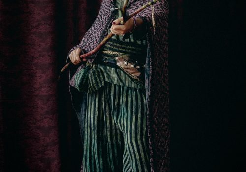 A hooded, cloaked figure stands ready for action in front of a burgundy backdrop, holding a forked wooden staff toward the viewer. The figure wears a handwoven cloak with a diamond weave pattern in a reddish-pruple and black textured patterning, with a bejeweled black clasp and a horizontal striped silk tunic in multiple hues of green and black, striped handwoven pants and waistband in black and green, with assorted woven patches at the waist. They also wear an iridescent leather fanny pack adorned with evil eye pendants and a bead-encrusted face veil with purple and green beaded fringe.