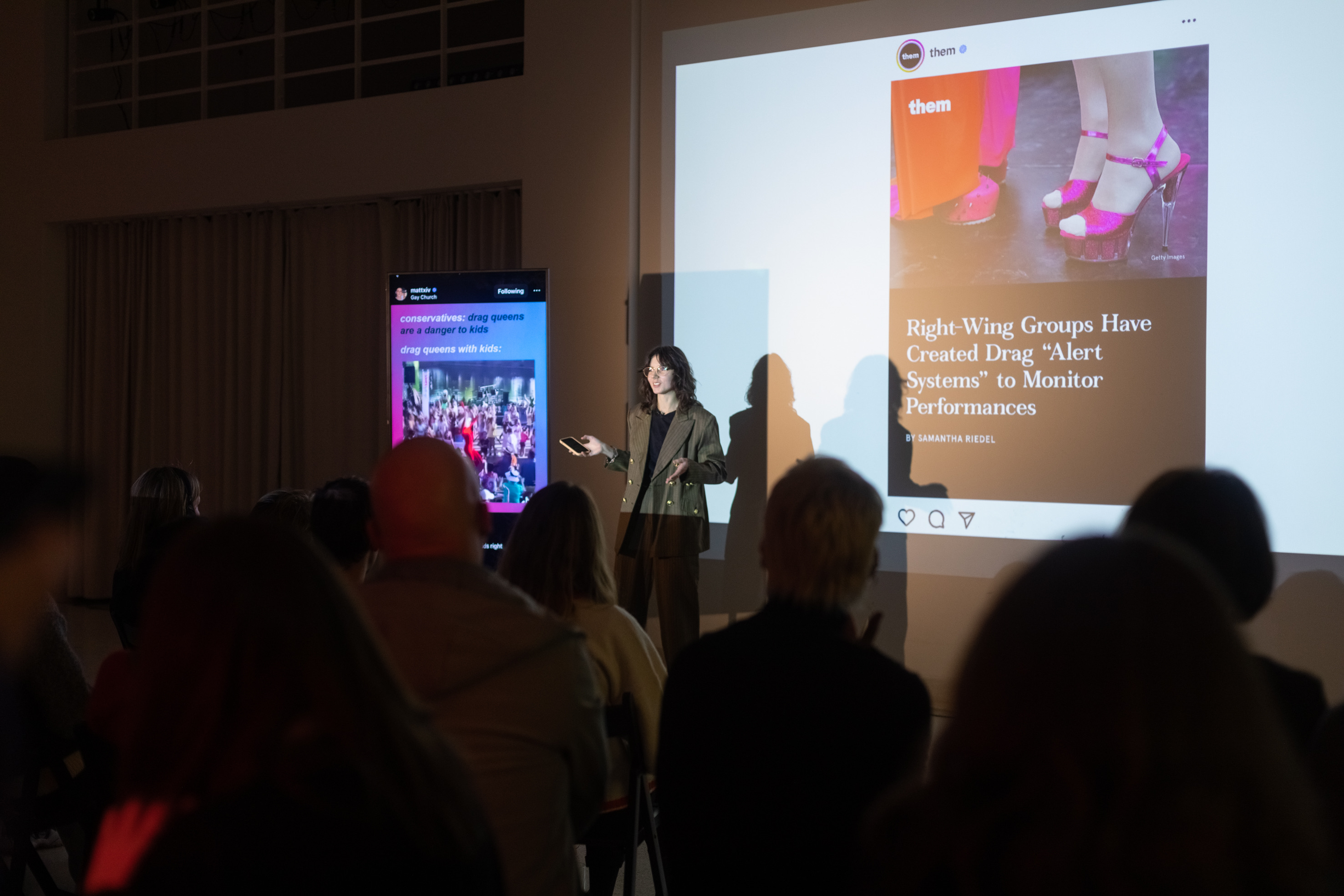 A person with phone in hand wearing a suit is giving a performative lecture in front of a crowd, a vertical TV is standing next to them and a presentation projection is behind them