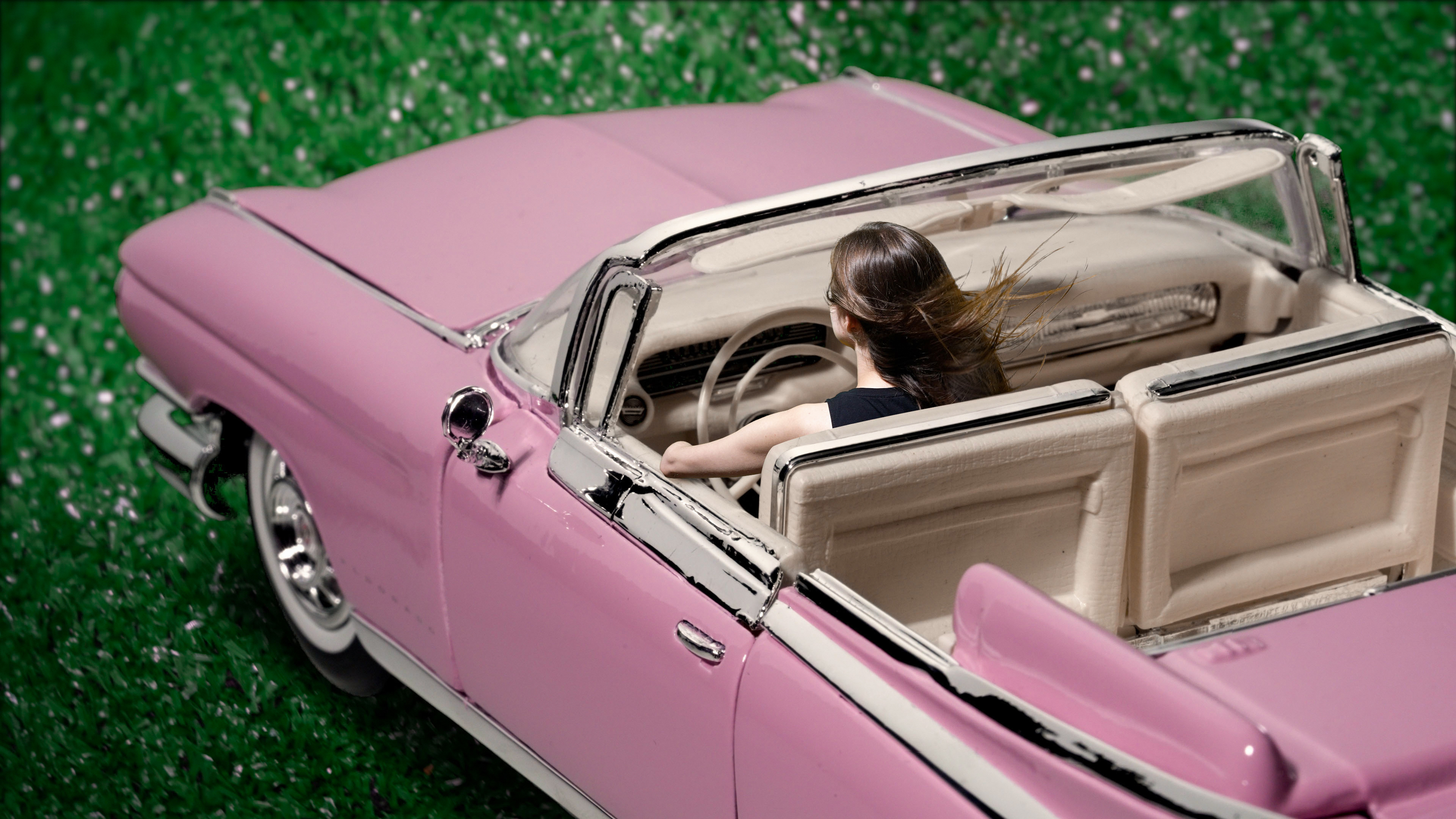 This image is a video still. It presents the front half of a toy pink convertible Cadillac, shot a bit from above, diagonally cutting through the frame, from the bottom right corner, heading towards the top left one, but not reaching it. The car is placed on a background made from fake green grass. In the car, at the steering wheel is a white woman with brown long hair flowing in the wind, wearing a black tank top. She is presented from the back, and is resting her left elbow on the door of the convertible. Her left arm ends right under her elbow.