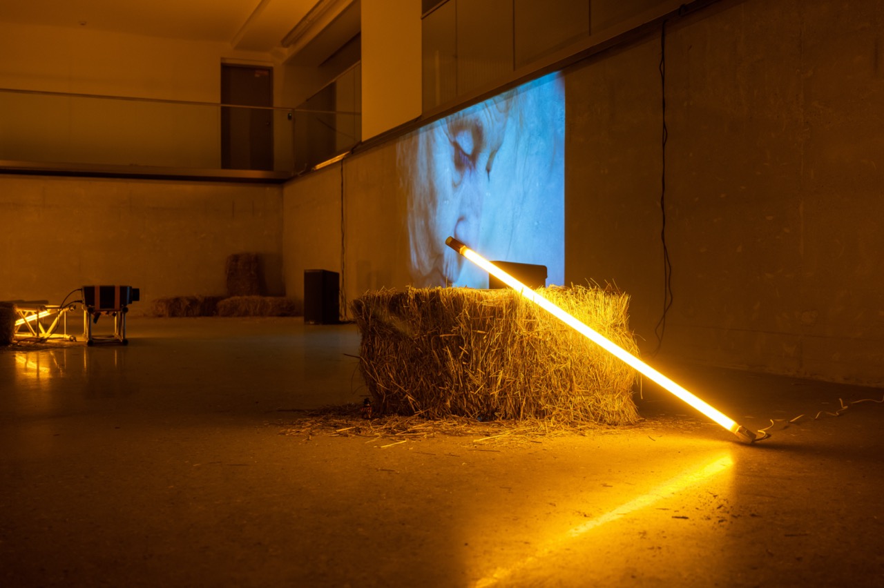 A bunndle of hay is placed on the ground with an led light covered in orange foil placed on top of it, in the background Joyful Flame is being projected on the wall