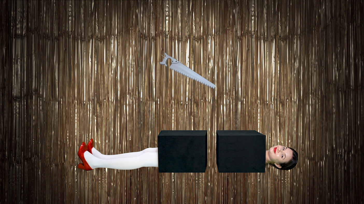 This image is a video still. The background of the image is filled with a gold festive curtain, made from individual strands of metallic ribbon. The middle of the frame is lit with round spotlight lighting. In the spotlight is a white woman laying down horizontally across the frame. She has white tights, red sequin shoes and red lips, her brown hair is in a bun. Her body is hidden in two separate black boxes that seem to have come from one longer box previously cut in half. As a result, her body appears to have been cut in half, most probably with the gray plastic-toy like saw hovering above her.